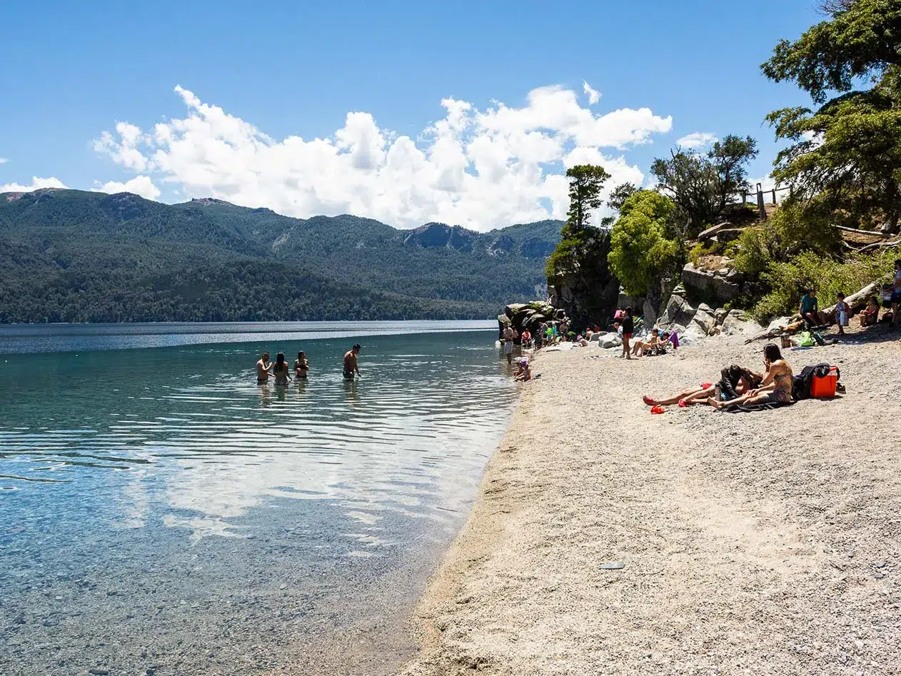 Playa de canto rodado con gente disfrutando del lago en verano.