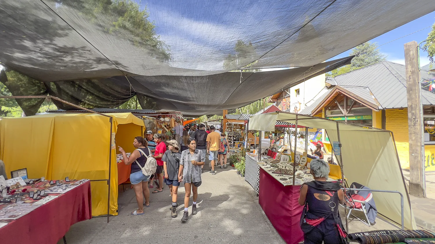 Feria artesanal de El Bolsón en la Plaza Pagano, con puestos de artesanías bajo media sombra y gente recorriendo.